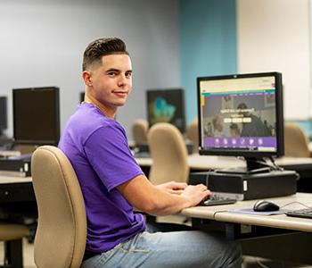 穿着紫色衬衫的学生坐在电脑前，看着身后的摄像头