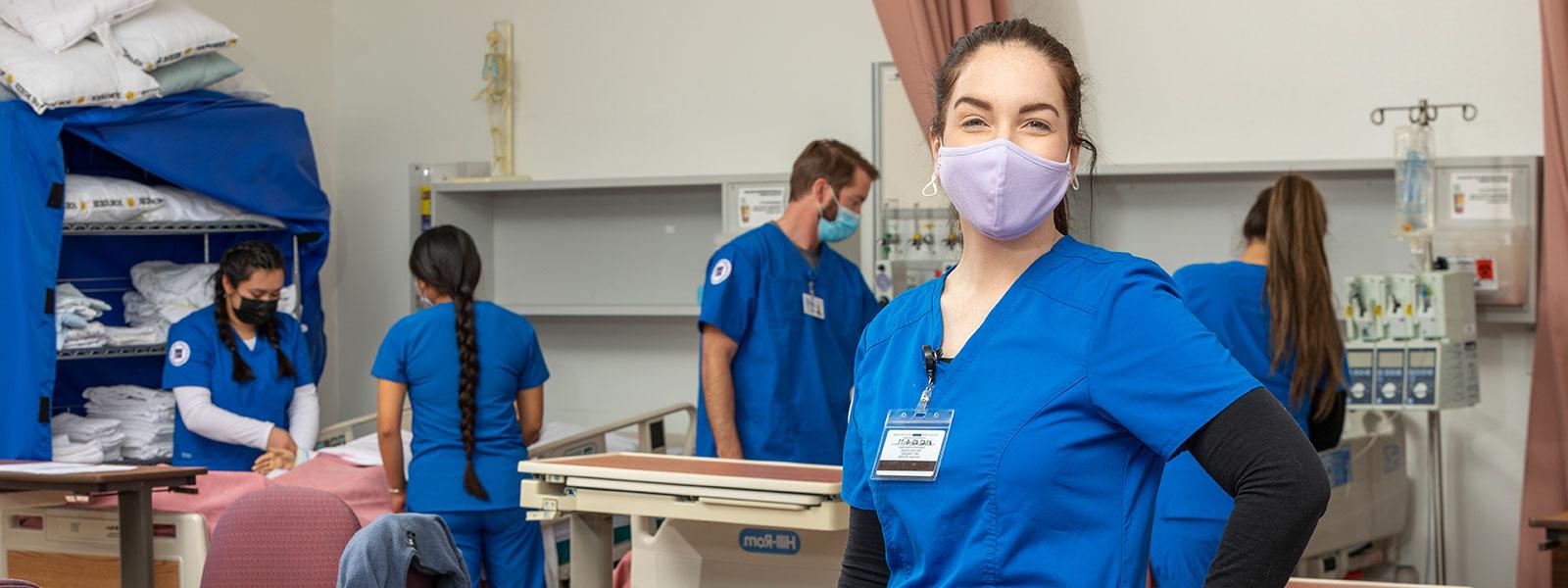 护士穿着工作服，戴着口罩看着镜头，而同学们在模拟实验室工作